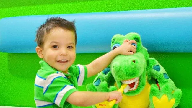 Young boy brushing teeth of a green stuffed dinosaur toy in a colorful room with blue and green decor.