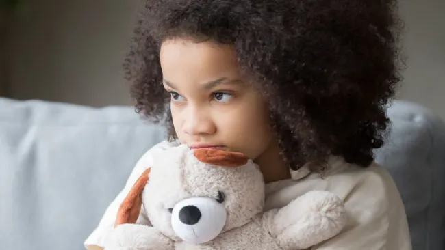 Thoughtful child with curly hair hugging a stuffed teddy bear while sitting on a sofa indoors