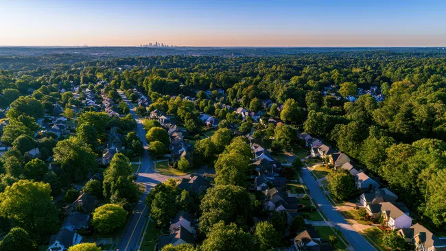 Aerial view of suburban neighborhood with tree-lined streets and distant city skyline under clear sky at sunset.