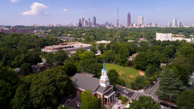 Aerial view of a city skyline with tall buildings, lush green trees, and a church with a steeple in the foreground.
