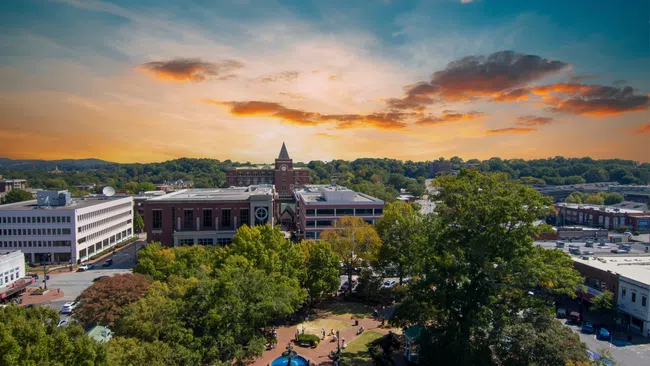 Aerial view of a town square with a central fountain, surrounded by trees and historic buildings at sunset.