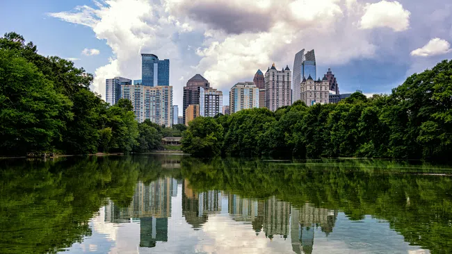 City skyline reflected in calm water with green trees and cloudy sky overhead