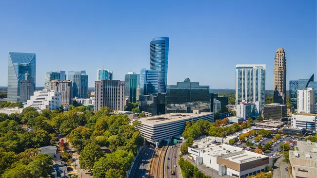 Aerial view of a modern city skyline with skyscrapers, green trees, and a railway track under a clear blue sky.