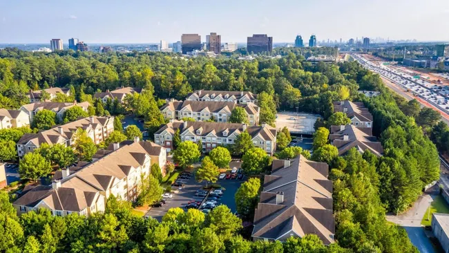 Aerial view of residential buildings surrounded by trees with a city skyline and highway in the background on a clear day