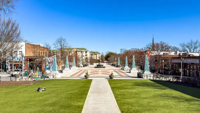 Wide pedestrian plaza with green lawns, geometric walkway, decorative towers, and clear blue sky in urban area.