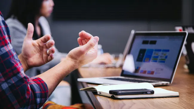Person gesturing with hands during a meeting with a laptop and notebook on the table in a modern office.