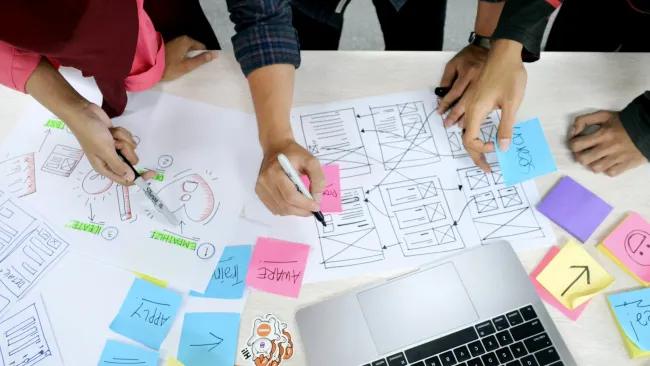Team collaborating on website design wireframes with markers, sticky notes, sketches, and laptop on a white table.