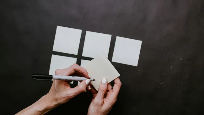 Hands writing on a sticky note with a marker on a black surface with four blank sticky notes above.