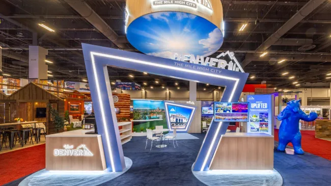 Modern Denver tourism booth with illuminated geometric frame, digital displays, and blue bear sculpture at expo
