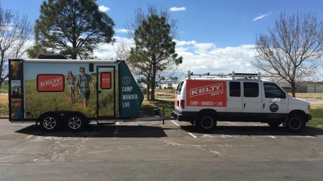White van towing trailer with Kelty outdoor brand logos parked near trees under a blue sky with clouds.