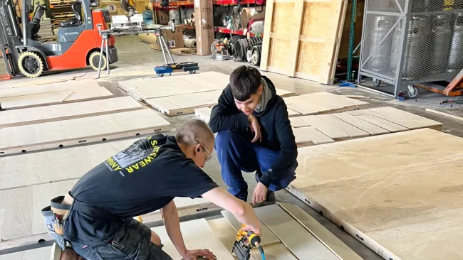 Two workers installing light-colored wooden floor panels in a workshop with tools and equipment nearby.