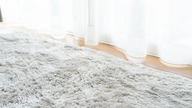 Soft fluffy white shag rug on light wooden floor in front of sheer white curtains.
