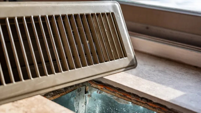 Close-up of a metal floor vent cover removed, showing the ductwork and window sill nearby in natural light