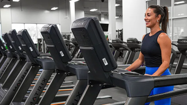 Woman in blue leggings and black tank top running on a treadmill in a modern gym with multiple Matrix treadmills.