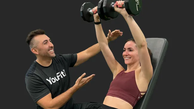 Female lifting dumbbells on incline bench with male personal trainer coaching and smiling