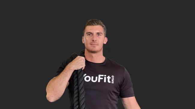 Man in black YouFit Gyms t-shirt holding black battle rope on dark background with confident expression.
