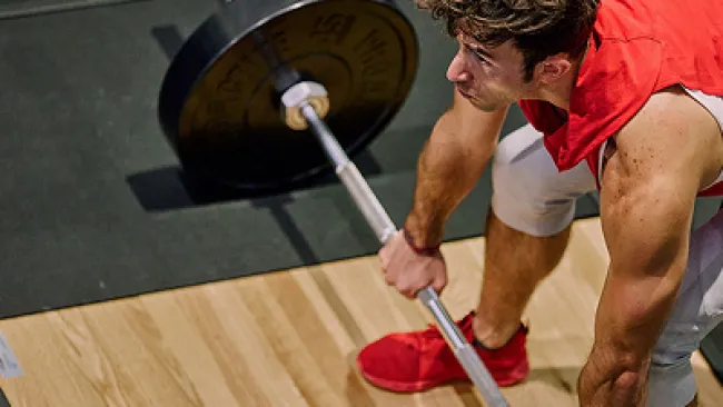 Man in red lifting barbell on wooden gym floor preparing for weightlifting exercise.