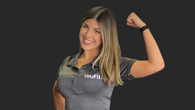 Smiling woman in gray YouFit shirt flexes arm muscle against dark background, showing strength and fitness.