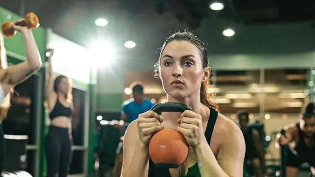 Focused woman doing kettlebell squats in a gym with other people exercising in the background.