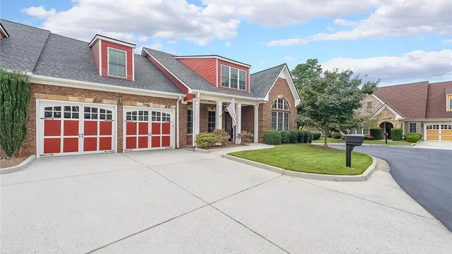 Suburban brick house with red accents, three-car garage, manicured lawn, and clear blue sky.