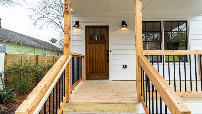 Front porch of a house with wooden railings, a wooden door, white siding, and outdoor wall lights.