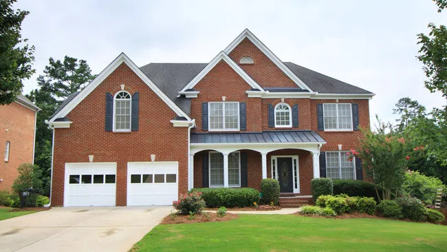 Large two-story brick house with black shutters, double garage, front porch, and green lawn on a clear day