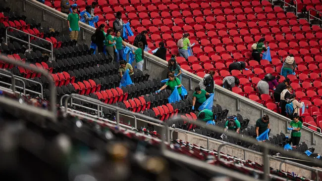 Volunteers in green shirts cleaning empty red and black stadium seats holding blue trash bags.