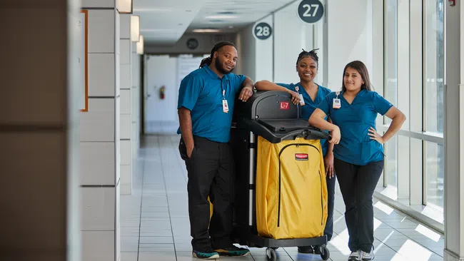 Three janitors in blue uniforms standing with a yellow cleaning cart in a bright hallway with numbered signs.