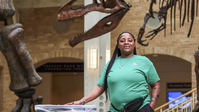 Janitor with green shirt and cleaning cart posing in museum with dinosaur skeleton display behind them.