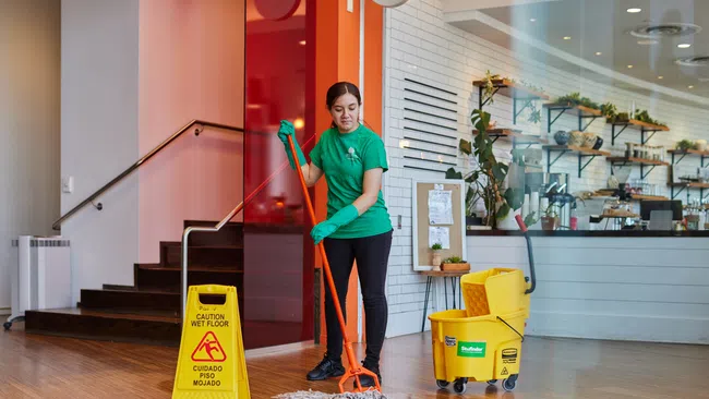 Woman in green shirt mopping wooden floor in commercial space with wet floor caution sign and yellow mop bucket.