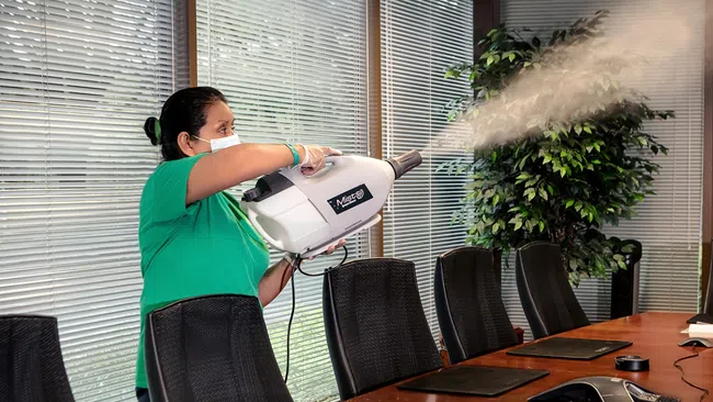 Person wearing mask spraying disinfectant fog in a modern office conference room with glass windows and chairs.