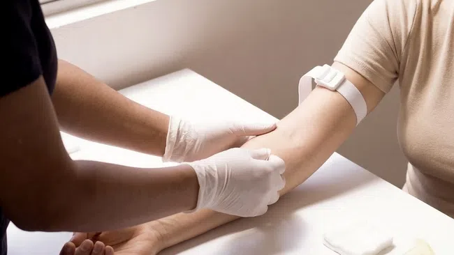 Medical professional wearing gloves preparing to draw blood from patient's arm with tourniquet applied.
