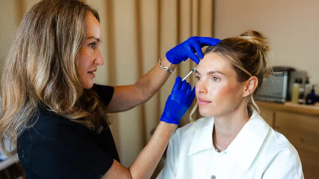 A cosmetologist wearing blue gloves applies facial injections to a woman in a white coat in a clinical setting.