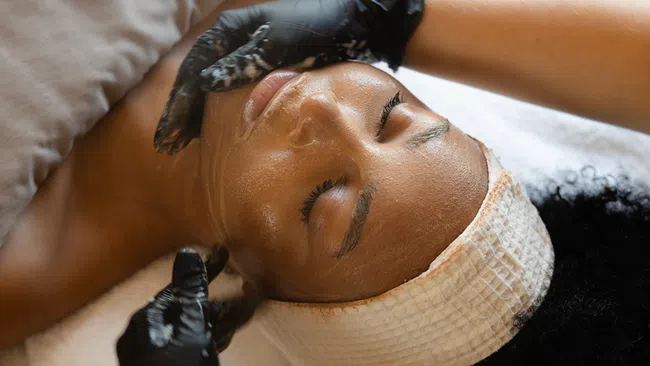 Woman receiving a facial treatment with gloves on in a spa setting, eyes closed and headband on.