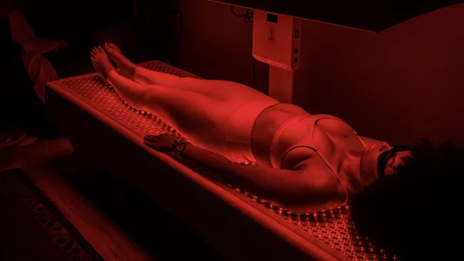 Woman in athletic wear lying on a bed under red light therapy panel in a dark room for wellness treatment.