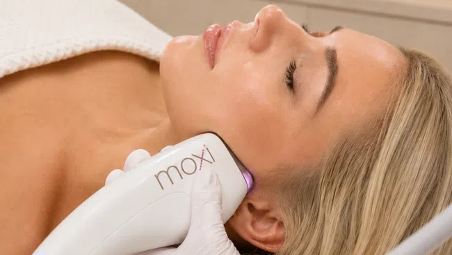 Woman receiving Moxi skin treatment with handheld device on her cheek in spa setting.