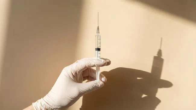 Hand wearing white glove holding syringe with shadow on beige wall background, medical injection concept