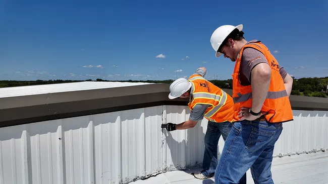 Two Mattco roofing technicians in orange safety vests inspecting a commercial metal roof edge and parapet wall on a clear day in Texas. One technician is standing and examining the flashing detail while the other crouches to assess the base of the wall. Mattco branding is visible on the standing technician vest.