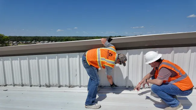 Two Mattco roofing technicians in orange safety vests inspecting a commercial metal roof edge and parapet wall on a clear day in Texas. One technician is standing and examining the flashing detail while the other crouches to assess the base of the wall. Mattco branding is visible on the standing technician's vest.