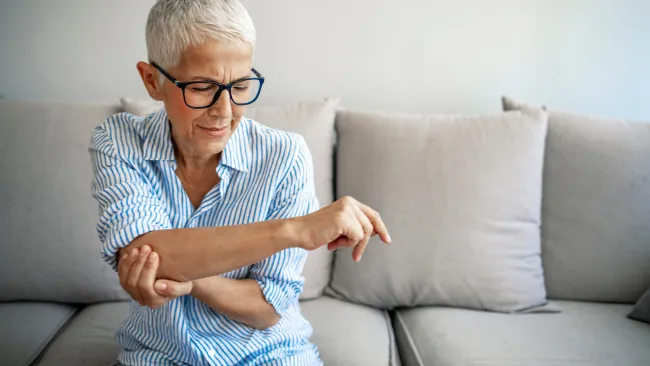 Senior woman with short gray hair and glasses sitting on a gray couch, holding her painful elbow.