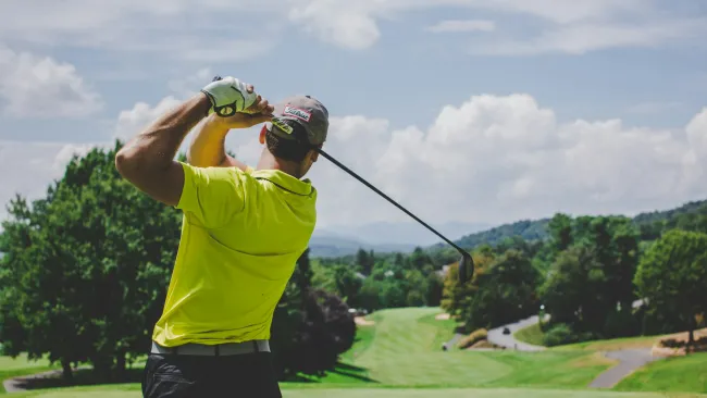Golfer in a yellow shirt swinging a golf club on a lush green course under a partly cloudy sky