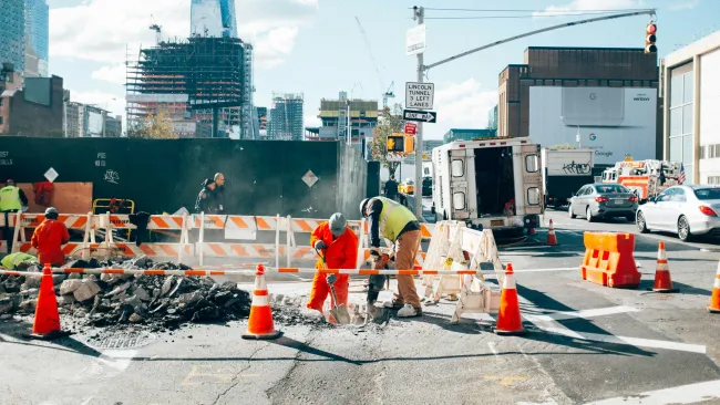 Construction workers repair urban road surrounded by traffic cones and city skyscrapers in daylight.