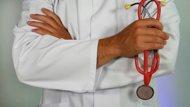 Doctor in white coat holding a red stethoscope with folded arms against light background