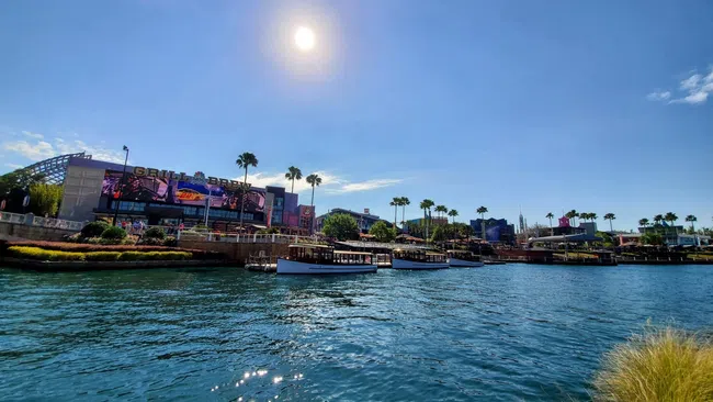 Sunny waterfront scene with boats docked near palm trees and buildings under a clear blue sky.