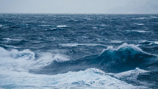 Stormy ocean waves with white foam under cloudy sky and distant foggy mountains in background