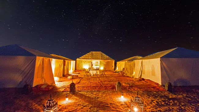 Desert night camp with illuminated tents, lanterns, carpets, and starry sky overhead highlighting a warm outdoor setting.