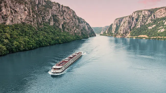 River cruise ship sailing through a scenic canyon with steep rocky cliffs and calm blue water under a pink sky.