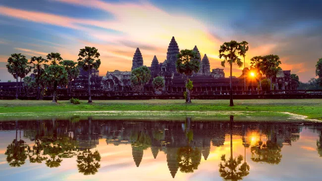 Sunset over Angkor Wat temple with palm trees reflected in calm water in Cambodia