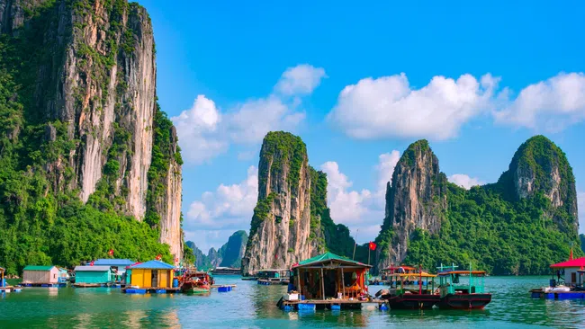 Floating houses and boats on calm water with towering limestone karsts under a partly cloudy blue sky.