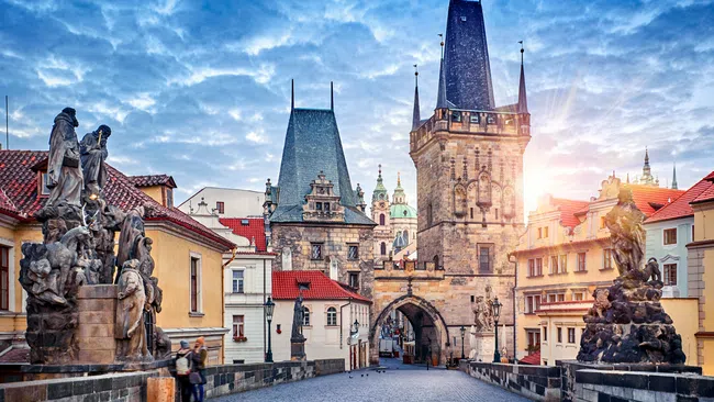 Early morning view of historic Charles Bridge with Gothic towers and statues in Prague under a partly cloudy sky.